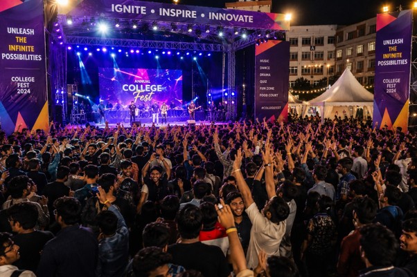 Wide shot of an energetic Indian college fest scene with students cheering, colorful stage lighting, banners, and a crowd, evening atmosphere, dynamic event photography style